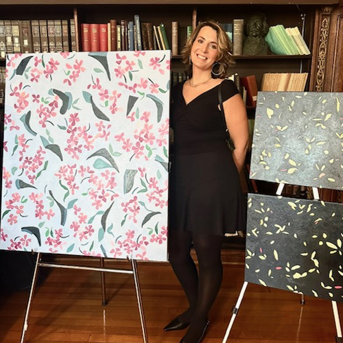 Ginger Mimmo Rohlfing stands next to a large painting of scattered green leaves and pink flowers, along with two smaller paintings with gray backgrounds and green leaves.